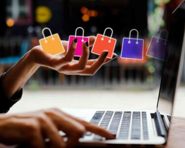 Dłoń nad klawiaturą laptopa, a nad nią unoszą się kolorowe ikony toreb zakupowych – symbol e-commerce i sprzedaży online.