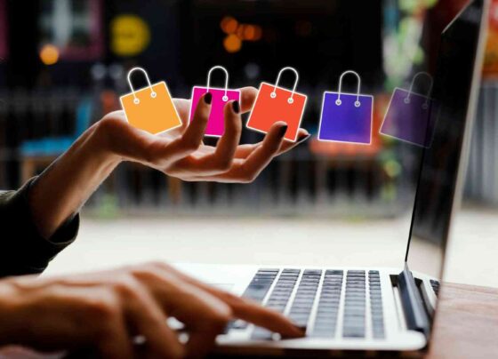 Dłoń nad klawiaturą laptopa, a nad nią unoszą się kolorowe ikony toreb zakupowych – symbol e-commerce i sprzedaży online.